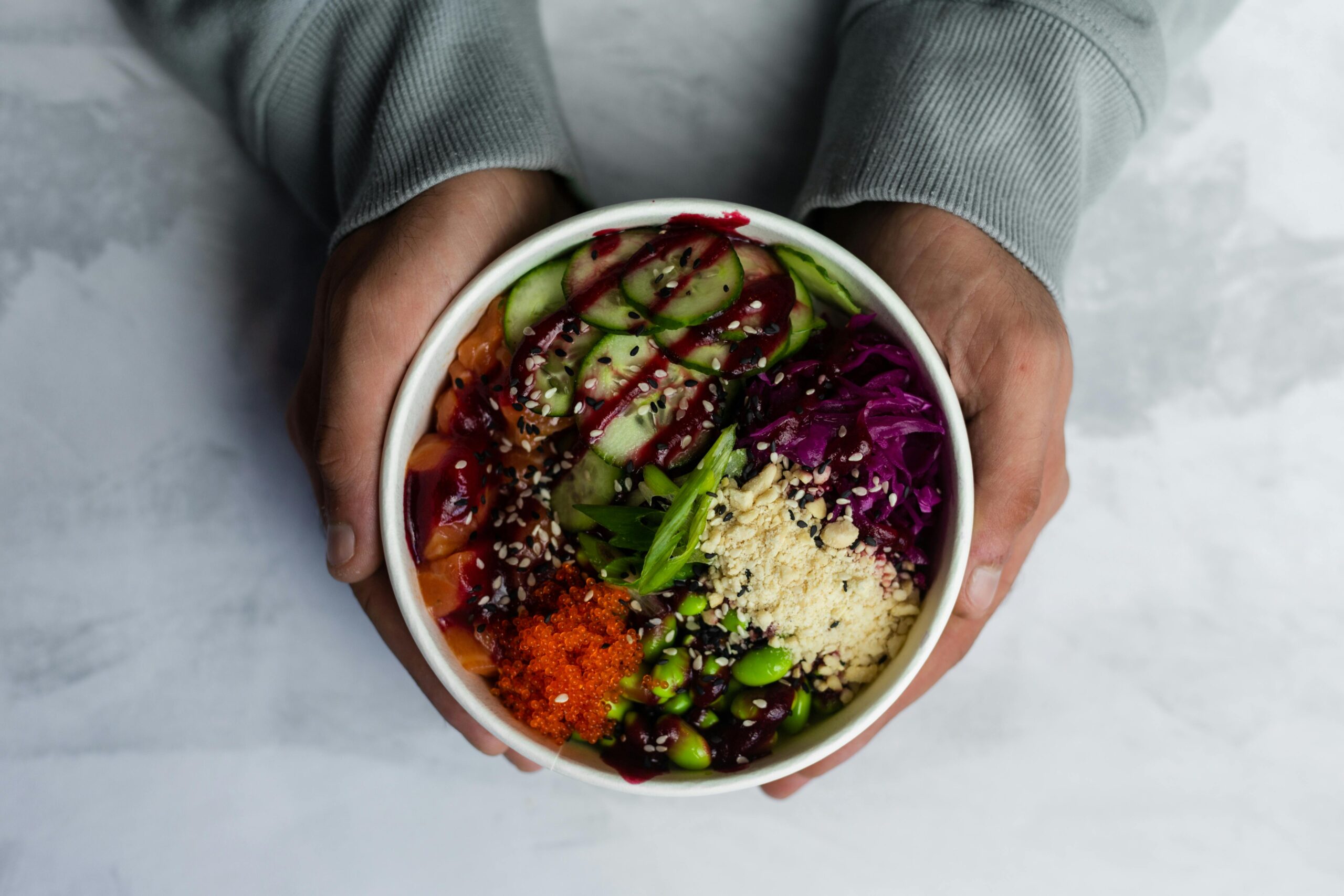 A vibrant poke bowl with fresh vegetables and toppings held in hands on a light background.